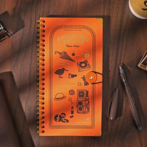May include: An orange spiral-bound notebook with a gold wire coil and a circular elastic closure. The cover features the text "COLLECTED THINGS" and "Paper Keeps" alongside various black line art illustrations including a camera, umbrella, and hat.
