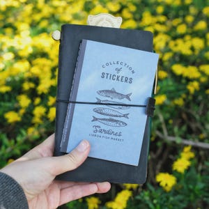 May include: A black leather journal with a light blue sticker book that reads "Collection of Stickers" and "Sardines Lisbon Fish Market." The journal is held together with a black elastic band and a gold-colored metal bookmark.
