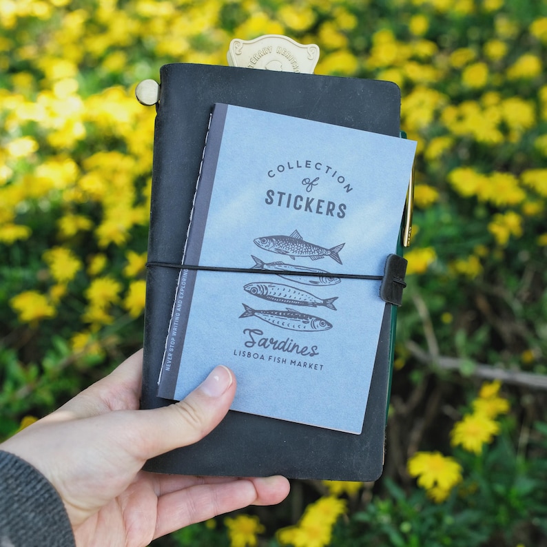 May include: A black leather journal with a light blue sticker book that reads "Collection of Stickers" and "Sardines Lisbon Fish Market." The journal is held together with a black elastic band and a gold-colored metal bookmark.