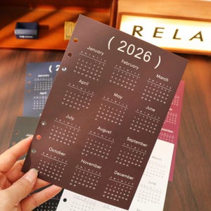 Pu&ograve; includere: Una pagina di calendario marrone del 2026 con testo bianco, con i mesi da gennaio a dicembre. Il calendario &egrave; perforato per la rilegatura. La parola "RELA" &egrave; visibile sullo sfondo.