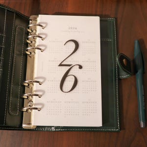 May include: Open dark green leather planner with a 2026 calendar insert. The calendar page features large black numbers and a monthly layout. A dark green pen rests beside the planner.