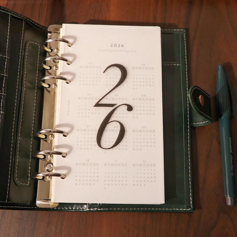 May include: Open dark green leather planner with a 2026 calendar insert. The calendar page features large black numbers and a monthly layout. A dark green pen rests beside the planner.