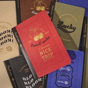 May include: A selection of travel notebooks in diverse colours, such as red, black, and blue. The red notebook displays gold foil text and a cherry illustration. Other notebooks feature phrases like "Lucky Every Day" and "Nice Trip."