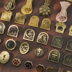 May include: A collection of metal pins and clips featuring various designs including hands, flowers, books, cats, and paw prints. The items are rendered in gold, bronze, and rose gold tones, showcasing intricate engraved details and dark accents.