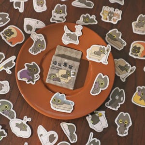 May include: A collection of cat stickers scattered on a brown plate and wooden surface. The stickers feature various poses of cartoon black cats, some with text in Chinese and English. A small box of stickers is also present, with the text "Mischievous black cat."