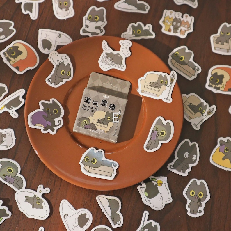 May include: A collection of cat stickers scattered on a brown plate and wooden surface. The stickers feature various poses of cartoon black cats, some with text in Chinese and English. A small box of stickers is also present, with the text "Mischievous black cat."
