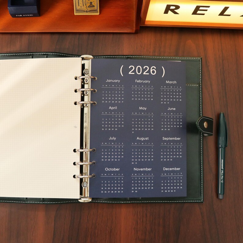 Pu&ograve; includere: Agenda in pelle verde scuro aperta con un inserto calendario 2026. L'agenda &egrave; aperta per rivelare il calendario e una pagina bianca a griglia. Una penna nera &egrave; appoggiata sulla superficie di legno.