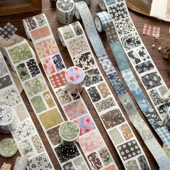 May include: Washi tape rolls featuring  and geometric patterns in green, pink, blue, and grey. The tape is unrolled, displaying the repeating designs. Several rolls are visible, showing the end designs. The background is a wooden surface.