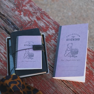 May include: Two light purple sticker collection notebooks featuring a black line drawing of a bear pushing a shopping cart. The text reads COLLECTION my STICKERS and Oops I bought stickers again. One notebook is smaller and attached to a black cover.