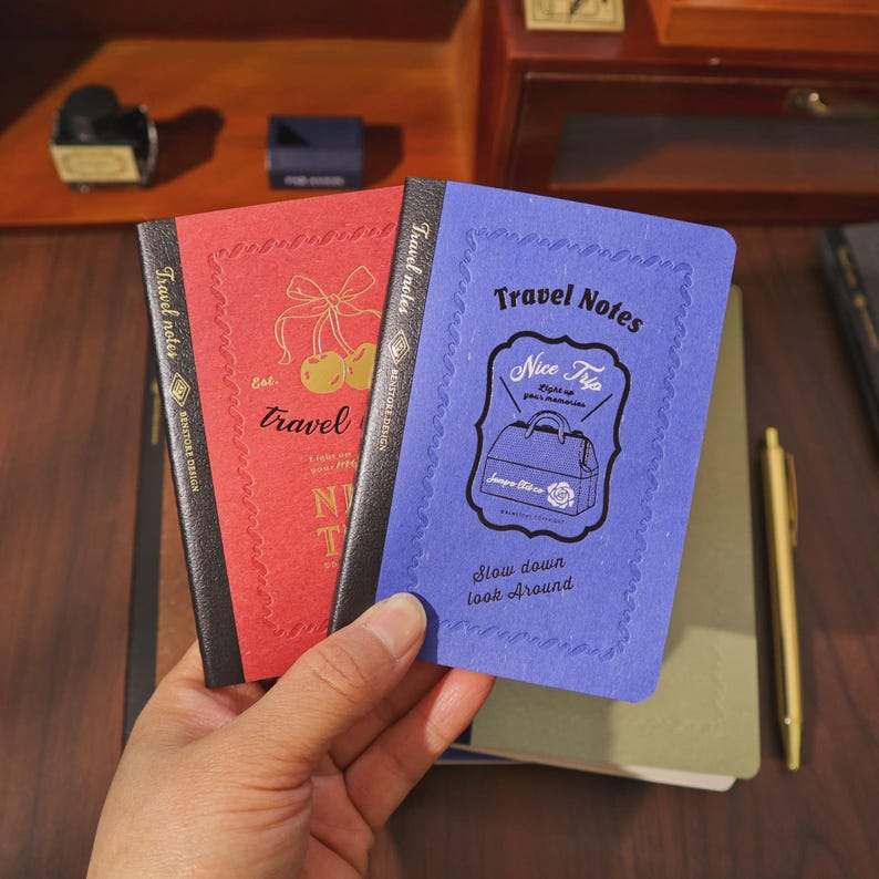 May include: Two small notebooks with black spines are held in a hand. One is red with gold text and a cherry design, the other is blue with a travel-themed graphic. Both say "Travel Notes".
