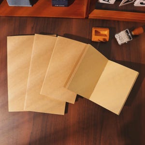 May include: Several open and closed blank kraft paper notebooks are arranged on a dark wooden surface. The notebooks are a light brown color, with a simple, minimalist design. A wooden stamp and ink pad are in the background.