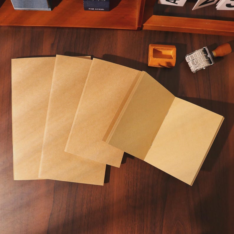May include: Several open and closed blank kraft paper notebooks are arranged on a dark wooden surface. The notebooks are a light brown color, with a simple, minimalist design. A wooden stamp and ink pad are in the background.