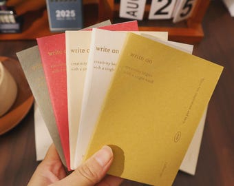 May include: A hand holding a fan of small notebooks in various colors: gray, red, cream, white, and mustard yellow. Each notebook has the words "write on" and "creativity begins with a single word" printed on the cover. A wooden calendar and a 2025 calendar are in the background.