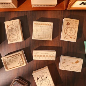 May include: A collection of wooden stamps with various designs, including a calendar, a to-do list, and a clock. The stamps feature cute cat illustrations and text such as "What Should I Do Today?" and "work challenge."