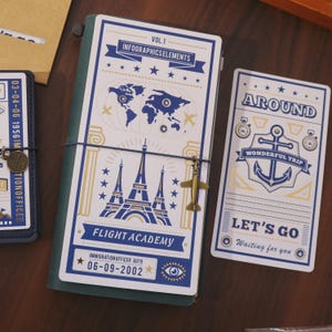 May include: A travel-themed journal with a blue and white cover featuring illustrations of the Eiffel Tower, a world map, and an airplane charm. The journal includes the text "FLIGHT ACADEMY" and "LET'S GO".