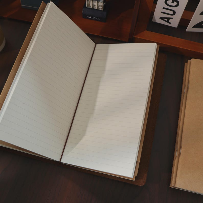 May include: An open, lined notebook with a brown leather cover. The pages are white with horizontal lines. A red elastic band holds the notebook closed. Another closed notebook is visible to the right.