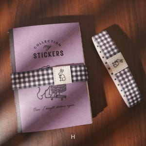May include: A purple notebook with the text "COLLECTION my STICKERS" and a black and white checkered band with a bunny graphic. A matching checkered ribbon with a bunny graphic is also present.