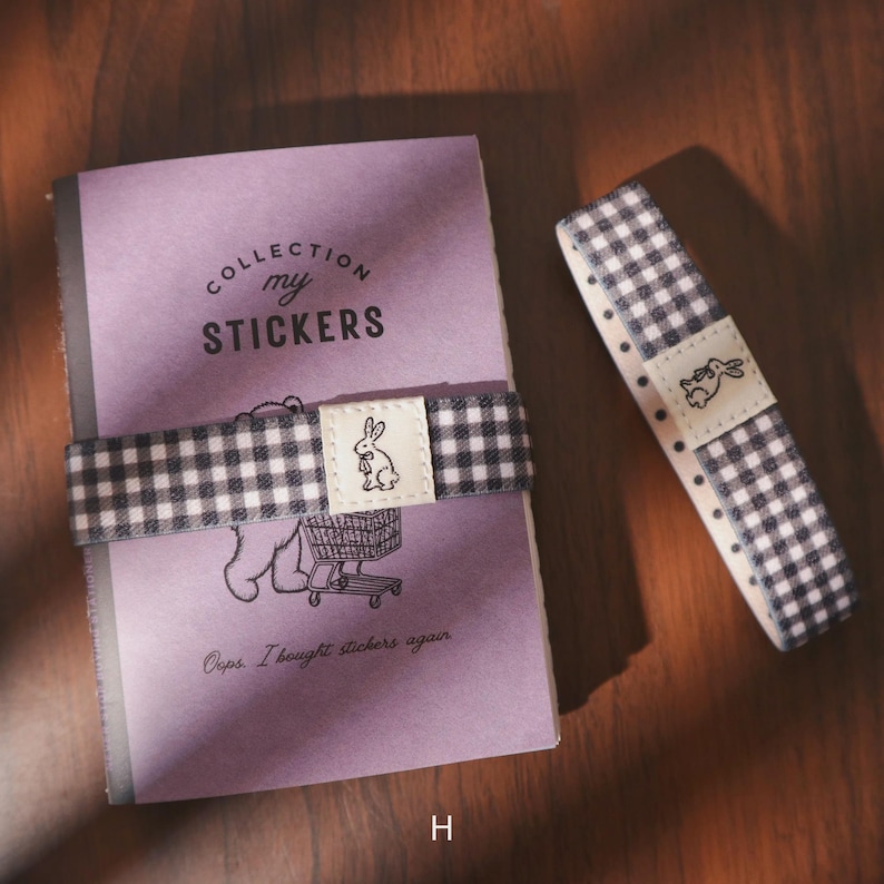 May include: A purple notebook with the text "COLLECTION my STICKERS" and a black and white checkered band with a bunny graphic. A matching checkered ribbon with a bunny graphic is also present.