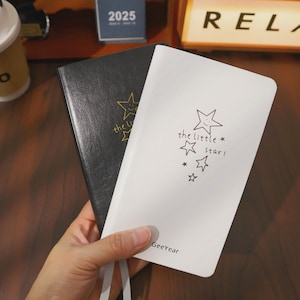 Puede incluir: Dos cuadernos, uno negro y otro blanco, se sostienen en una mano. El cuaderno blanco presenta un diseño de estrella con el texto "the little star!". El cuaderno negro tiene un diseño de estrella dorada. La marca "GeeYear" es visible.