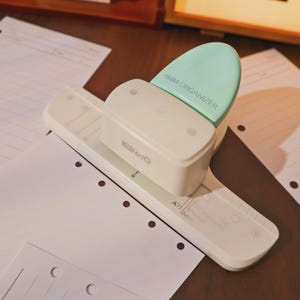 Könnte beinhalten: Ein weißer und mintgrüner KW-trio Papierlocher mit dem Text "4MM ORGANIZER" und "KW-trio" aufgedruckt. Der Locher befindet sich über einem weißen Blatt Papier mit vorgestanzten Löchern. Der Locher ist für A7 Mini-Papier konzipiert.