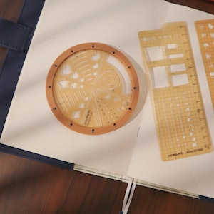 Könnte beinhalten: Ein Set von Schreibwaren, darunter eine beige kreisförmige Schablone mit verschiedenen Formen und ein Lineal mit dem Text "KOKUYO". Die Schablone hat einen braunen Rand und liegt auf einem Notizbuch mit Punkten.