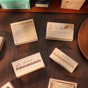 May include: A collection of wooden stamps with various designs. One stamp features a calendar layout, while others display weekly schedules and cute illustrations. The stamps are made of light-colored wood and are ideal for journaling and crafting.