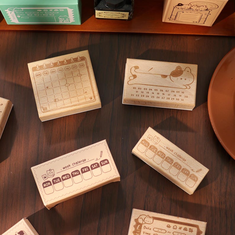 May include: A collection of wooden stamps with various designs. One stamp features a calendar layout, while others display weekly schedules and cute illustrations. The stamps are made of light-colored wood and are ideal for journaling and crafting.