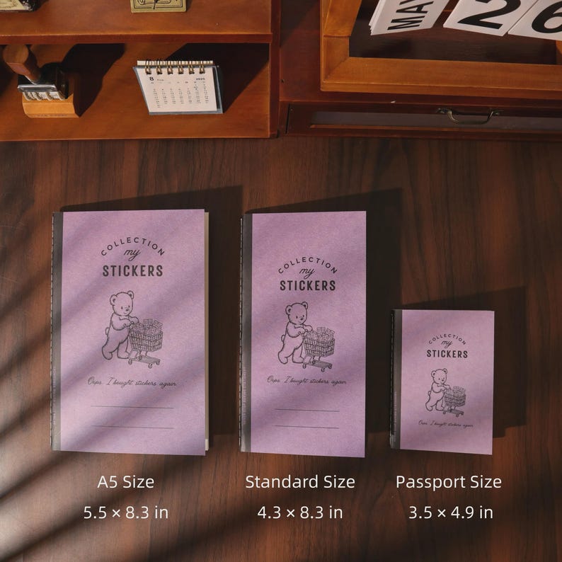 May include: Three purple sticker collection notebooks in A5, standard, and passport sizes. Each notebook features a black line drawing of a bear pushing a shopping cart, the text "COLLECTION my STICKERS", and "Oops I bought stickers again."
