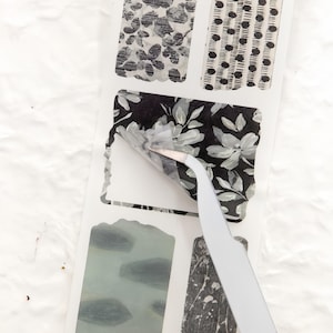 May include: A sheet of decorative stickers with various black and white patterns. One sticker is being peeled back with tweezers, revealing a floral design. Other patterns include abstract shapes and dotted lines.