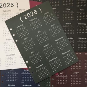 Pu&ograve; includere: Una collezione di calendari annuali del 2026. I calendari sono stampati su vari tipi di carta colorata, tra cui bianco, nero e blu scuro. Ogni pagina del calendario mostra i mesi dell'anno con i giorni della settimana. I calendari sono perforati per l'uso in raccoglitori.