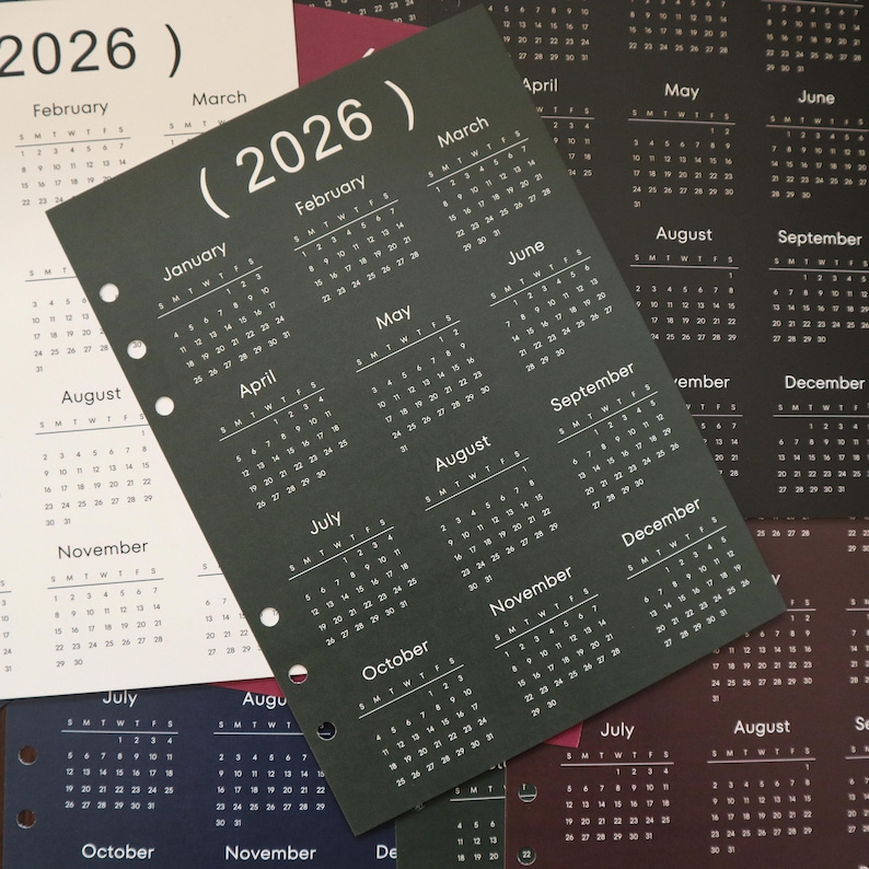 Pu&ograve; includere: Una collezione di calendari annuali del 2026. I calendari sono stampati su vari tipi di carta colorata, tra cui bianco, nero e blu scuro. Ogni pagina del calendario mostra i mesi dell'anno con i giorni della settimana. I calendari sono perforati per l'uso in raccoglitori.