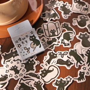 Op de afbeelding: Een verzameling zwart-witte kattenstickers verspreid op een houten oppervlak. Een kleine, lichtblauwe doos met tekst en een kattenillustratie is ook aanwezig. De stickers tonen verschillende kattenposes en -ontwerpen.