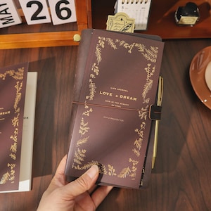Puede incluir: Un diario de cuero marrón oscuro con un borde botánico de lámina dorada y el texto "LIFE JOURNAL LOVE & DREAM LOVE IS THE BEAUTY OF SOUL Live a beautiful life." El diario está asegurado con una banda elástica marrón e incluye un bolígrafo dorado.
