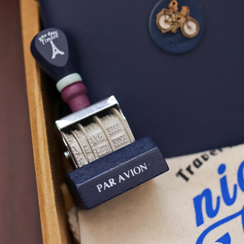 Op de afbeelding: Een marineblauwe en paarse datumstempel met de Eiffeltoren op de handgreep. De stempel leest "PAR AVION" en heeft een roterend datumwiel. Een fietspen is zichtbaar op de achtergrond.