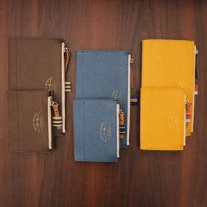 Puede incluir: Una colección de estuches y cuadernos con cremallera en varios tamaños y colores, incluyendo marrón, azul y amarillo. Cada artículo presenta una cremallera dorada y una lengüeta de cuero. Los artículos están dispuestos sobre una superficie de madera oscura. Medidas en centímetros.