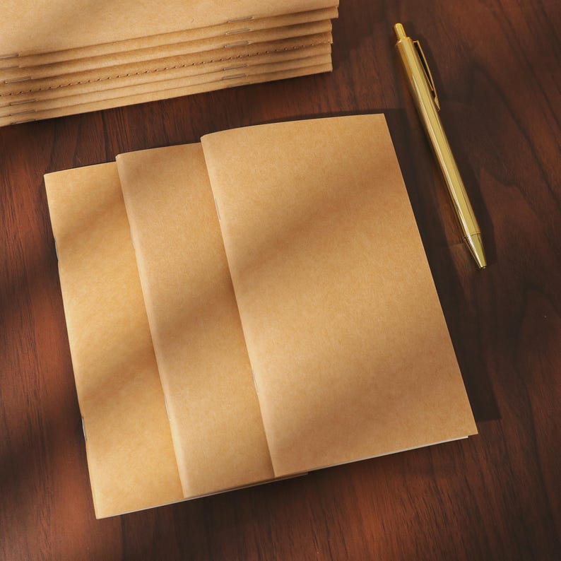 May include: A stack of kraft paper notebooks with three open, lying flat on a dark wood surface. A gold pen rests nearby. The notebooks are a light brown color.