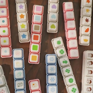 May include: A selection of small, square stamps in various colours and designs. The stamps feature shapes such as stars, hearts, and clovers, along with other cute illustrations. The stamps are arranged on a brown surface.