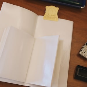 May include: Two open white notebooks with blank pages. A gold-colored metal clip with a decorative scalloped edge and embossed text is attached to the top of the larger notebook. The notebooks are positioned on a flat surface with a small black object.