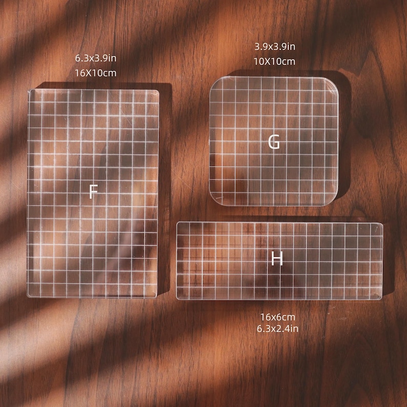 Puede incluir: Tres bloques de estampado acr&iacute;licos transparentes con patrones de cuadr&iacute;cula y las letras F, G y H. Los bloques son rectangulares y cuadrados, con medidas en cm y pulgadas. Los bloques est&aacute;n sobre una superficie de madera.