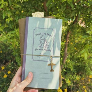 Può includere: Un diario con una copertina azzurra con il testo "Trust The Seeds, You Are Planting" e un'illustrazione di orso. Il diario è rilegato con un elastico marrone e ha un piccolo ciondolo a forma di aeroplano. Lo sfondo è un ambiente verde e frondoso.