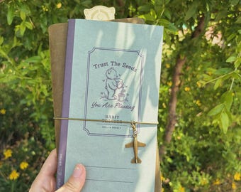 Może przedstawiać: Dziennik z jasnoniebieską okładką z napisem "Trust The Seeds, You Are Planting" i ilustracją misia. Dziennik jest oprawiony brązową gumką i ma mały samolotowy urok. Tło to zielone, liściaste otoczenie.