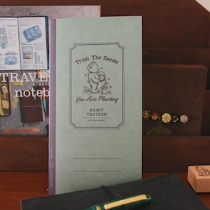 May include: A light green habit tracker notebook with the text "Trust The Seeds You Are Planting". The cover features a bear watering plants. The notebook is open, revealing lined pages, and is surrounded by other stationery items.