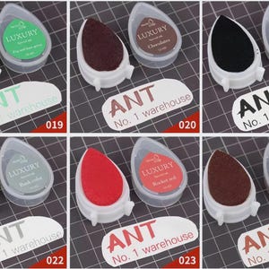May include: A collection of six ink pads in various colors, including green, brown, black, gray, and red. Each pad is in a white teardrop-shaped holder with a clear lid. The text "ANT No. 1 warehouse" is visible.