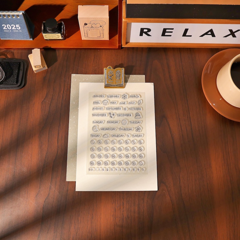 May include: A clear stamp set with months, days, and numbers for creating custom calendars. The stamp set is held in place by a gold-colored clip. The word "RELAX" is visible in the background.
