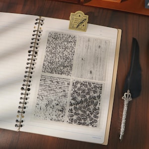 May include: A clear stamp set with four different patterns: a swirling design, wood grain, brick, and leaves. The stamps are on a notebook page with a gold-colored pen holder and a decorative quill pen.