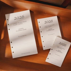 May include: Three white agenda diary refill pages for the year 2026, with the text "Week on Two Pages Diary Refill" and "Agenda Planning Diary". The pages have pre-punched holes for binding and are arranged on a wooden surface.