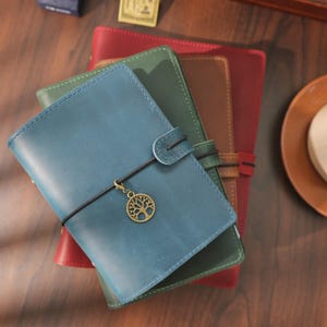Könnte beinhalten: Ein Stapel aus drei Lederjournalen in Blau, Grün und Rot. Das oberste blaue Journal hat ein schwarzes elastisches Verschlussband mit einem runden, bronzefarbenen Baum-Anhänger. Jedes Journal hat eine glatte, matte Oberfläche und eine rechteckige Form.