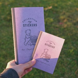 May include: Two purple sticker collection booklets featuring a dark brown illustration of a bear pushing a shopping cart filled with sticker packs. The text reads COLLECTION my STICKERS and Oops. I bought stickers again. The booklets have dark spines.
