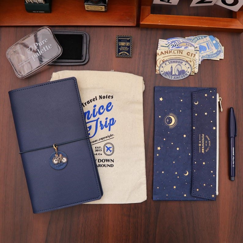 Op de afbeelding: Een verzameling reisbenodigdheden op een houten oppervlak. Bevat een marineblauw leren notitieboek met een fietsbedel, een canvas tas met tekst, een notitieboek met sterrenhemelthema en een zwarte pen. Ook stickers en een transparant palet.