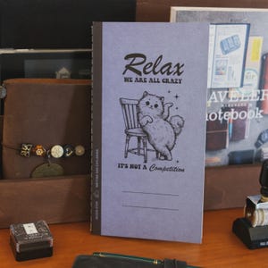May include: A lilac notebook featuring the word "Relax" and the phrase "WE ARE ALL CRAZY" in black script. A cartoon cat is illustrated next to a chair. The phrase "IT'S NOT A Competition" is below the cat illustration.